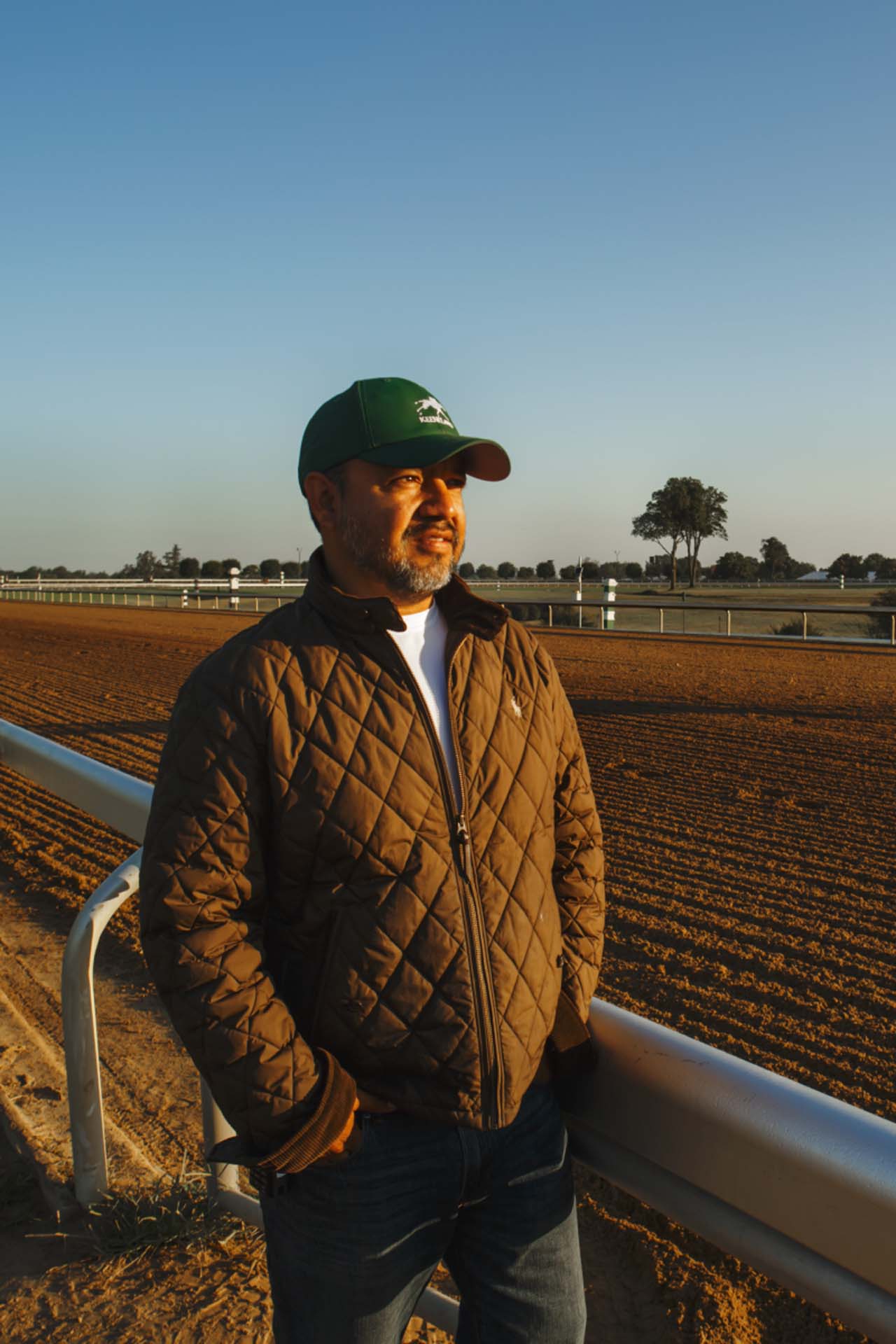 A photo of Alfredo Laureano looking off into the distance while standing by the dirt track at sunset. He is an older Hispanic man with graying chin stubble. He is wearing a green Hoofstrike-brand hat and a brown Hoofstrike-brand jacket overtop a white shirt.