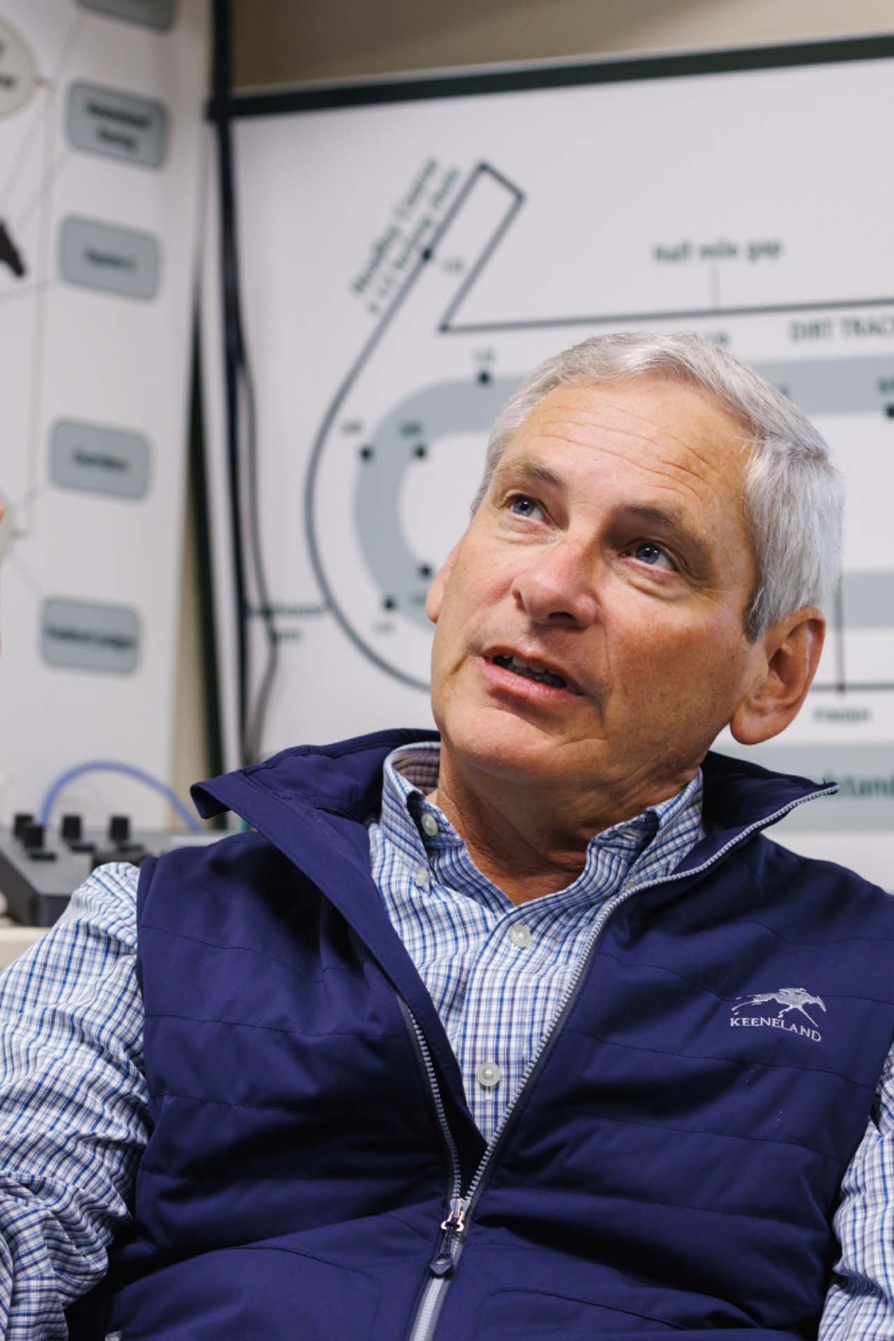 An up-close photo of Dr. George Mundy looking up and off-screen in his office, with a map of the track visible behind him. He is an older White man with short silver hair. He is wearing a navy Hoofstrike-brand vest over a white and blue checkered shirt.